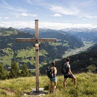 © www.fabianfranzferdinand.com Zwei Wanderer vor Gipfelkreuz mit der Inschrift 'Ehre sei Gott in der Höhe' und Blick auf Berge und Täler | © www.fabianfranzferdinand.com